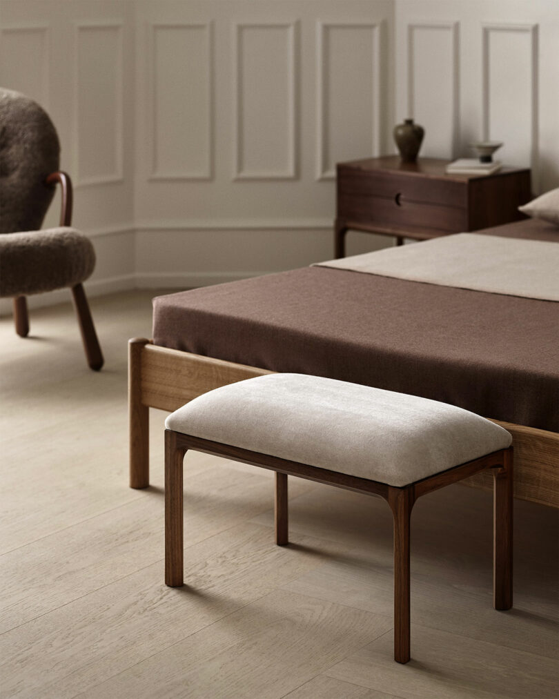 Minimalist bedroom featuring a wooden bed, brown bedding, upholstered bench, and an Entre Kai Kristiansen armchair beside the nightstand, with a decorative vase on light wood flooring.
