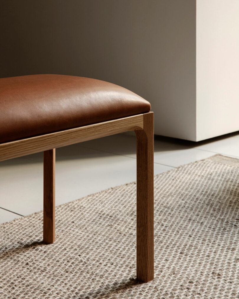 A close-up of an Entre wooden chair or bench with a brown leather seat on a textured beige rug, next to a tiled floor and a white wall.
