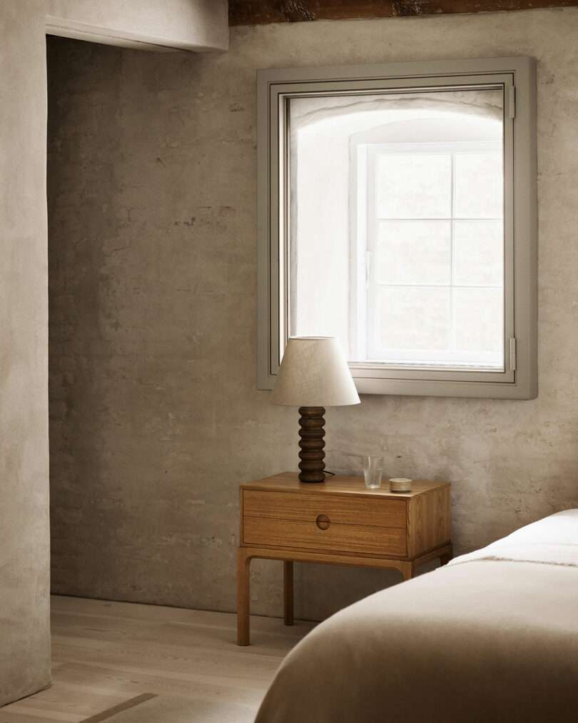 A minimalist bedroom with a wooden Entre nightstand, a lamp, a glass of water, and a window framed by textured, light-colored walls.