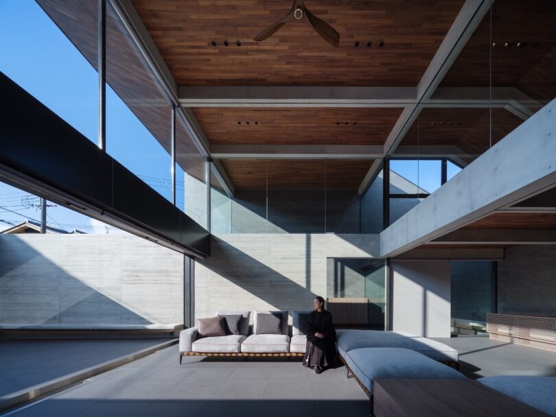 A person sits on a modern sofa in a spacious, minimalist living room with high ceilings, large glass windows, and wooden accents.