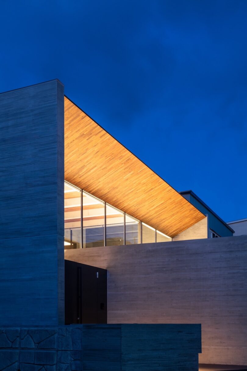 Modern building with concrete walls and large glass windows, featuring a wooden ceiling illuminated by warm lights against a deep blue evening sky.