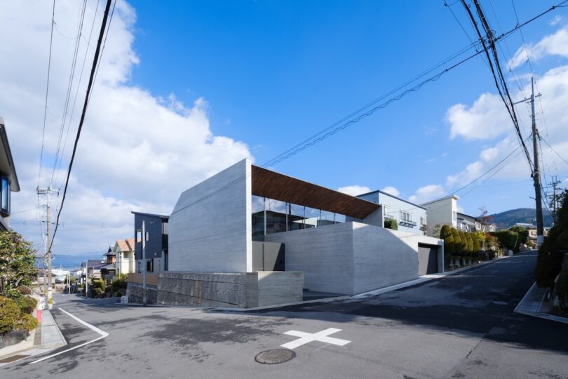 Modern concrete building with large windows situated on a street corner, surrounded by residential houses and overhead power lines under a blue sky with scattered clouds.