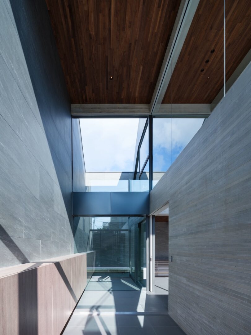 Modern interior hallway with high wood ceiling, large glass windows, and grey stone walls, featuring a glass balcony and natural light streaming in.