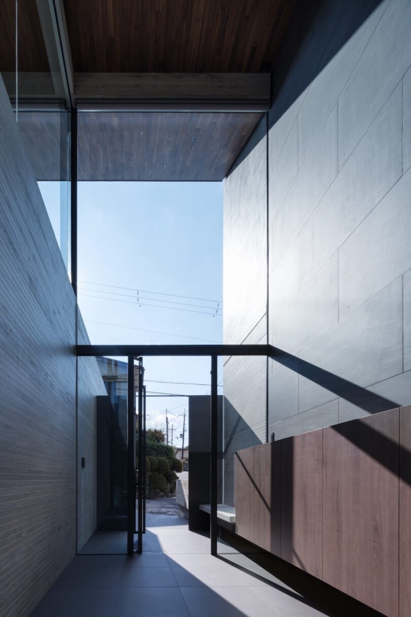 A modern entryway with floor-to-ceiling glass walls, wooden ceiling, tiled floor, and minimalist cabinetry, leading to an outdoor area with visible power lines and blue sky.