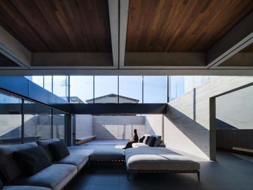A modern living room with large windows, minimalist grey furniture, wood ceiling, and an outdoor view; a person sits alone on a bench in sunlight.