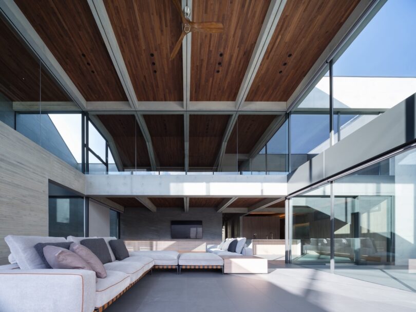 Modern living room with glass walls, wooden ceiling, grey flooring, and contemporary sofas, featuring an open, airy layout with ample natural light.
