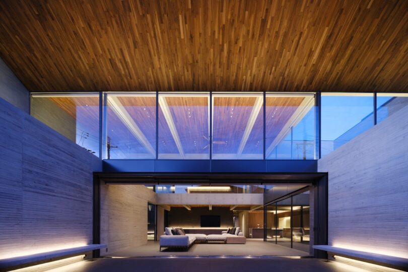 Modern living area with floor-to-ceiling glass windows, wood ceiling, concrete walls, and minimalist sofas, seen from an open courtyard at dusk.