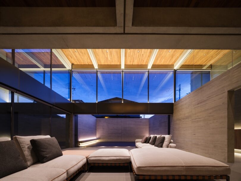 Modern living room with large glass windows, minimalist furniture, concrete walls, and a wooden ceiling, illuminated by natural evening light.