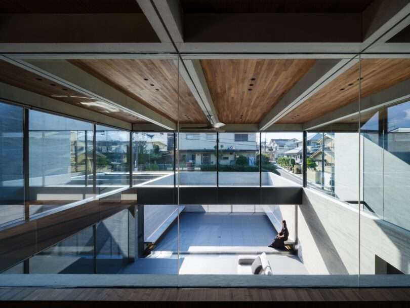 A person sits on a bench in a spacious, modern building featuring large glass windows, exposed concrete walls, and wooden ceilings with natural light streaming in.