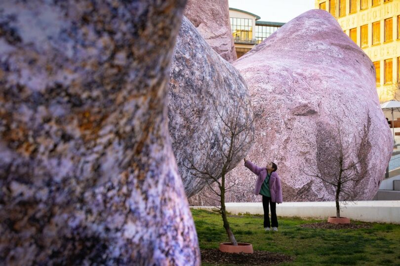A person stands among large, textured, purple-hued rock sculptures and small leafless trees in an outdoor urban area