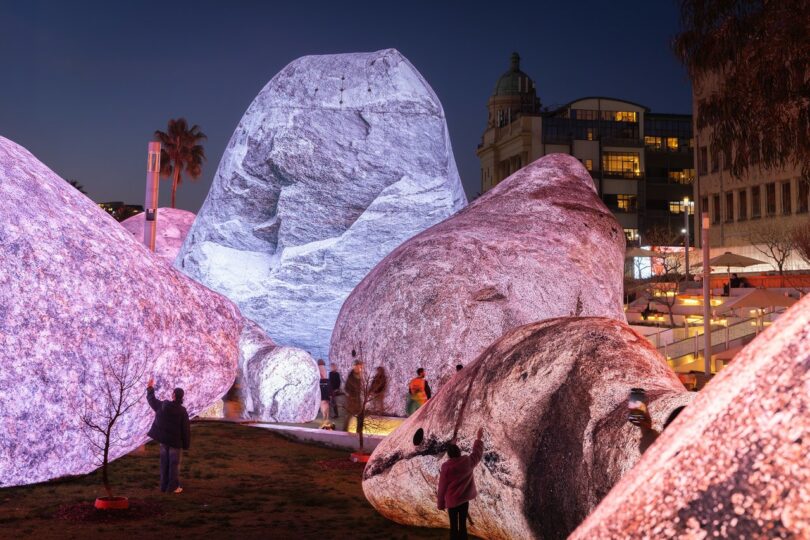 Several large boulders illuminated in pink and white light, with people standing and walking around them in an urban outdoor setting at dusk