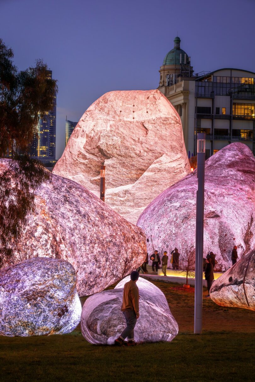 Several large boulders illuminated in pink and white light, with people standing and walking around them in an urban outdoor setting at dusk