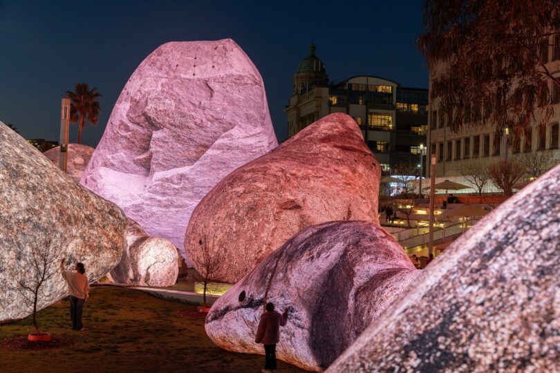 Several large boulders illuminated in pink and white light, with people standing and walking around them in an urban outdoor setting at dusk