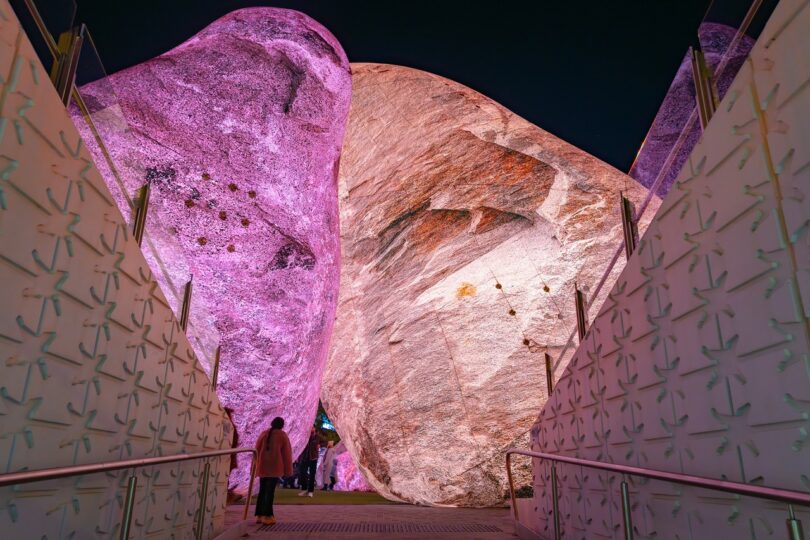 Several large boulders illuminated in pink and white light, with people standing and walking around them in an urban outdoor setting at dusk