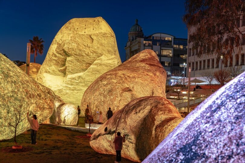 Large illuminated boulders are displayed outdoors at night, with several people walking and standing nearby; buildings and palm trees are visible in the background