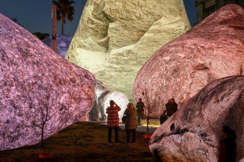 Several people stand among large, illuminated boulders outdoors at night, some taking photos; the rocks are lit with different colors, and small trees are visible nearby