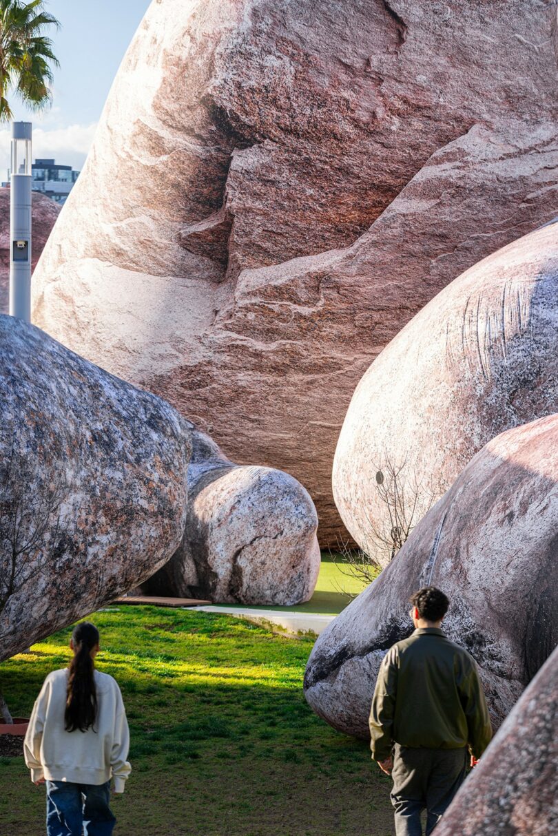 Two people walk on a path surrounded by large, realistic rock sculptures in an outdoor setting