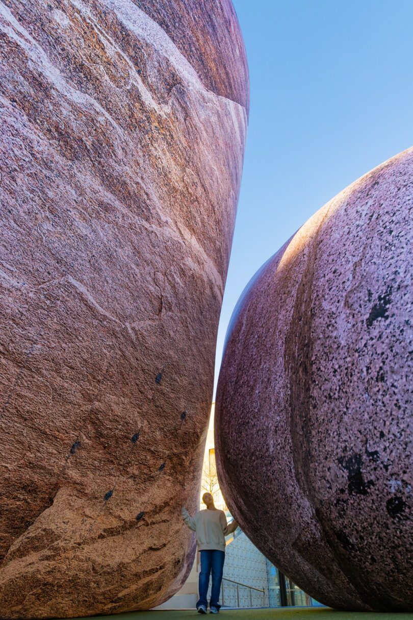 A person stands between two massive, smooth boulders with a clear blue sky above