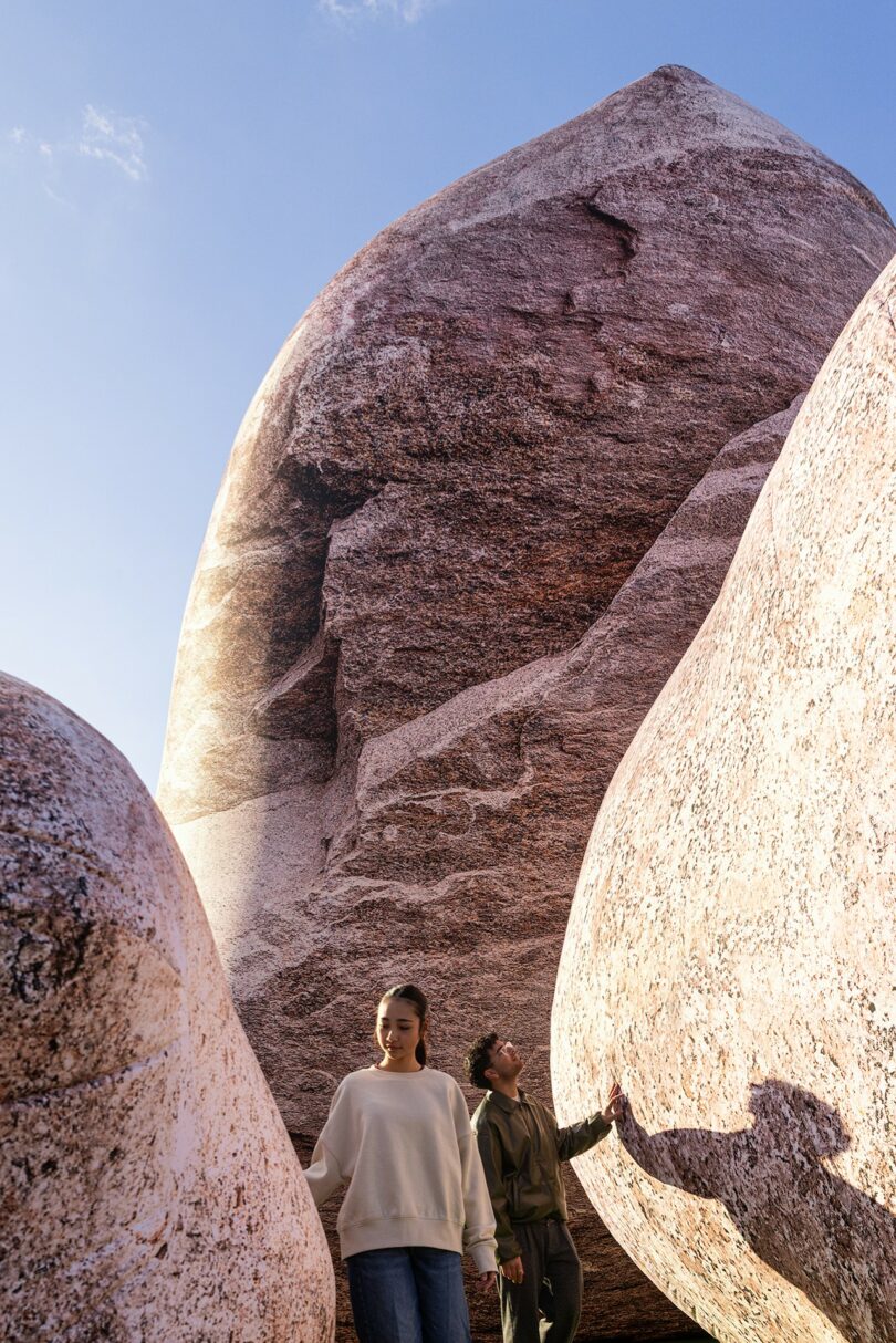 Two people stand among large reddish-brown boulders under a clear blue sky, with one person looking up and the other looking down