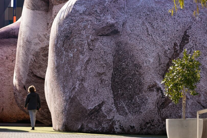 A person walks beside a large, textured sculpture resembling a stone figure, with a small potted tree nearby on a paved surface