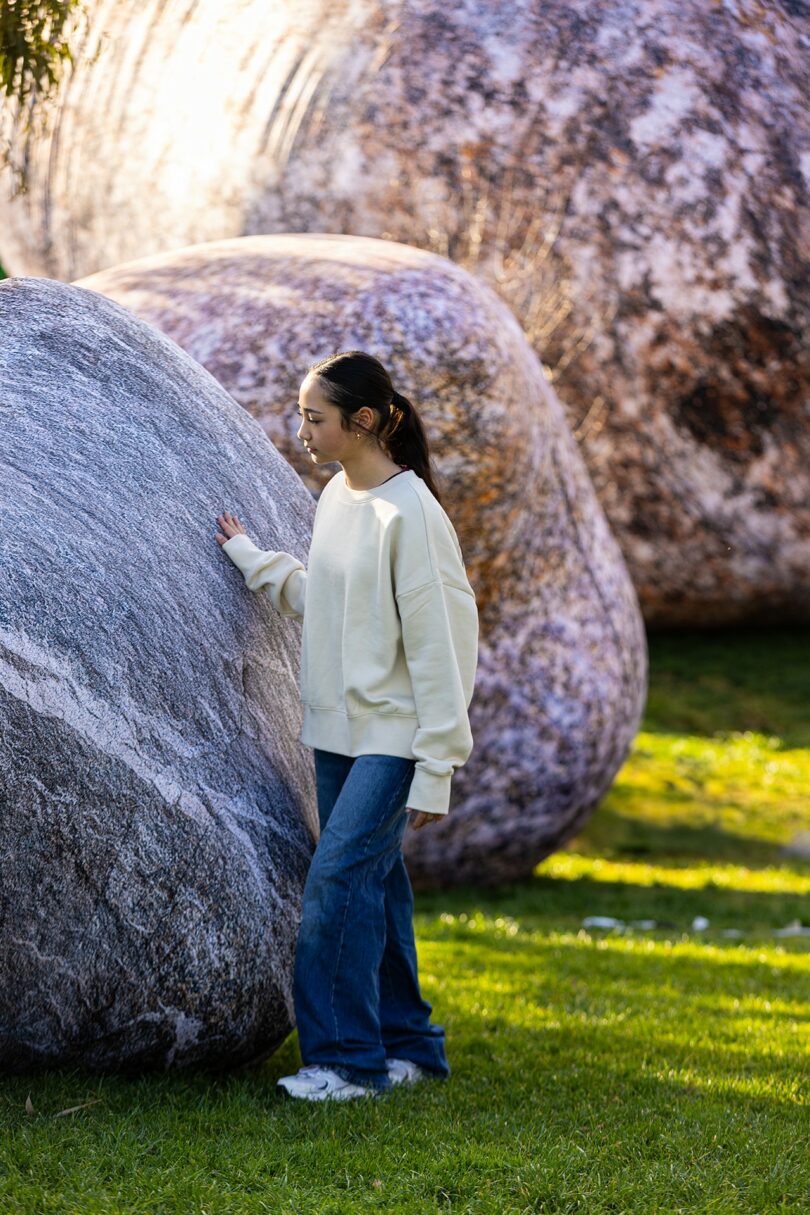 A person wearing a cream sweater and blue jeans touches a large rock while standing on green grass, with more large rocks in the background