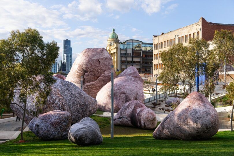 Large pink boulders scattered across a grassy urban park with people walking and running, modern and older buildings in the background
