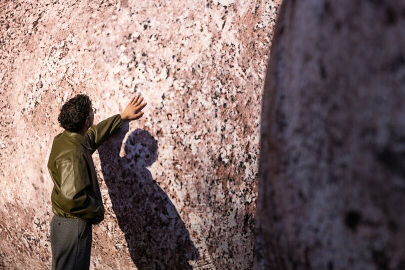 A person wearing a green jacket touches a large, textured rock surface, casting a shadow on it