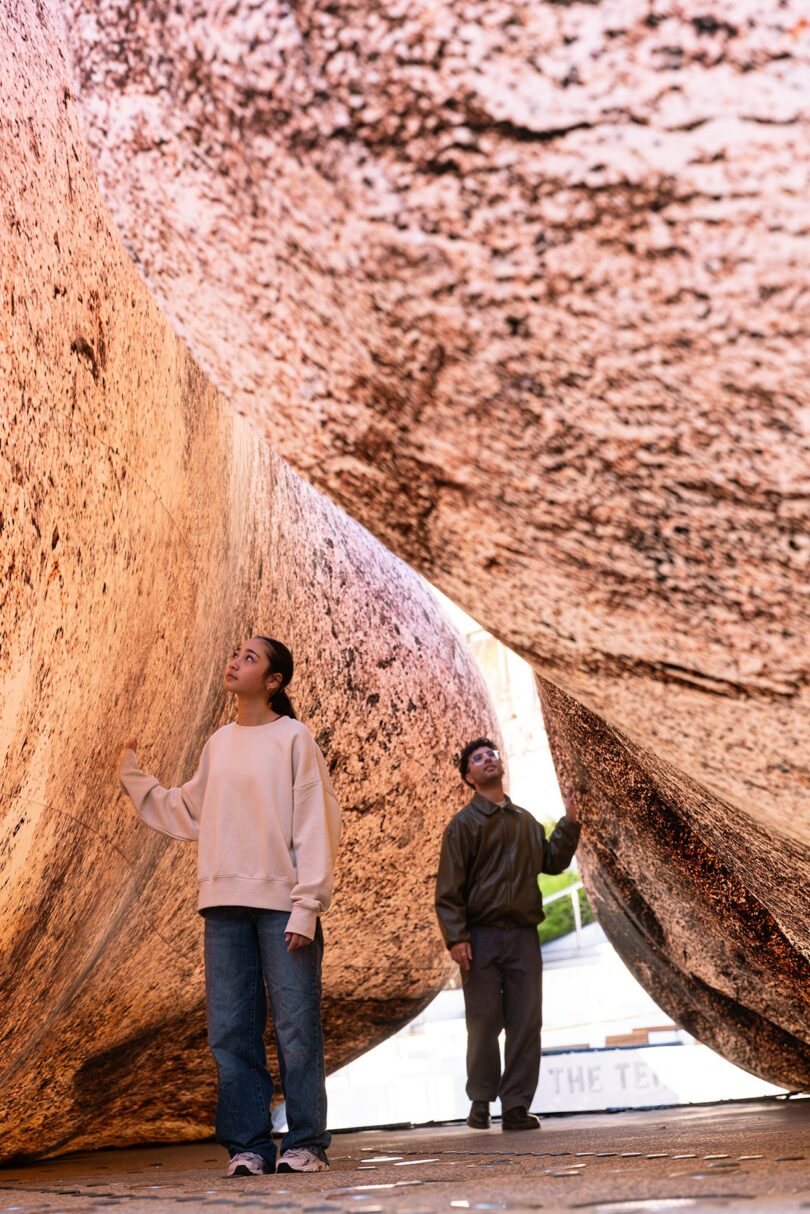 Two people stand and look up while walking between large, textured stone slabs in an outdoor setting