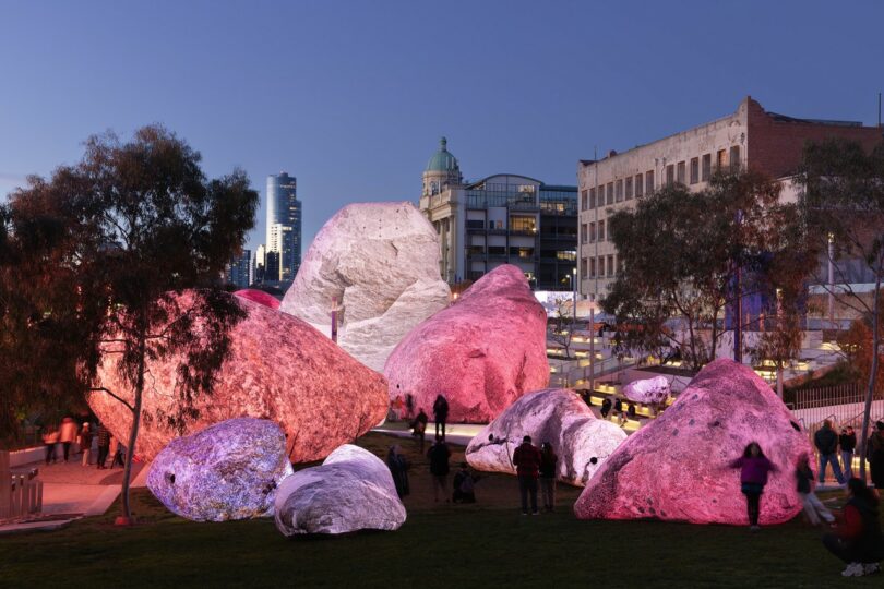 Large artificial boulders illuminated with pink and purple lights are scattered in an urban park at dusk, with people walking among them and city buildings in the background