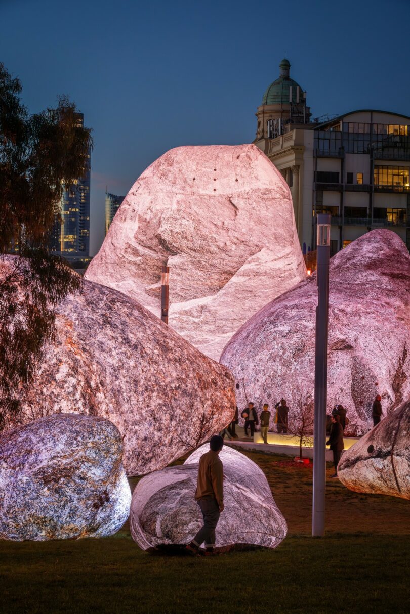 People stand among large, illuminated artificial boulders in an outdoor urban area at dusk, with buildings visible in the background