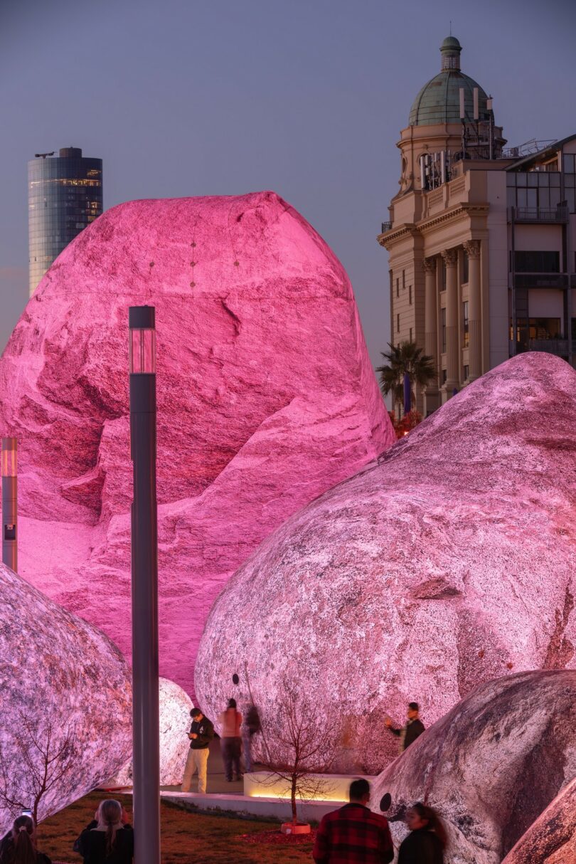 Large boulders illuminated with pink light, surrounded by people, with a historic building and a modern skyscraper in the background at dusk