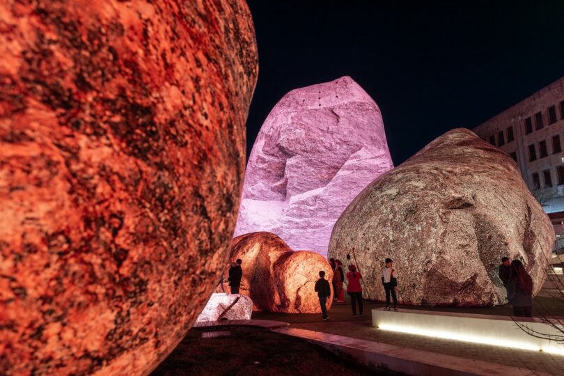 People stand near large illuminated boulders at night, with the rocks glowing in shades of red and purple under artificial lighting