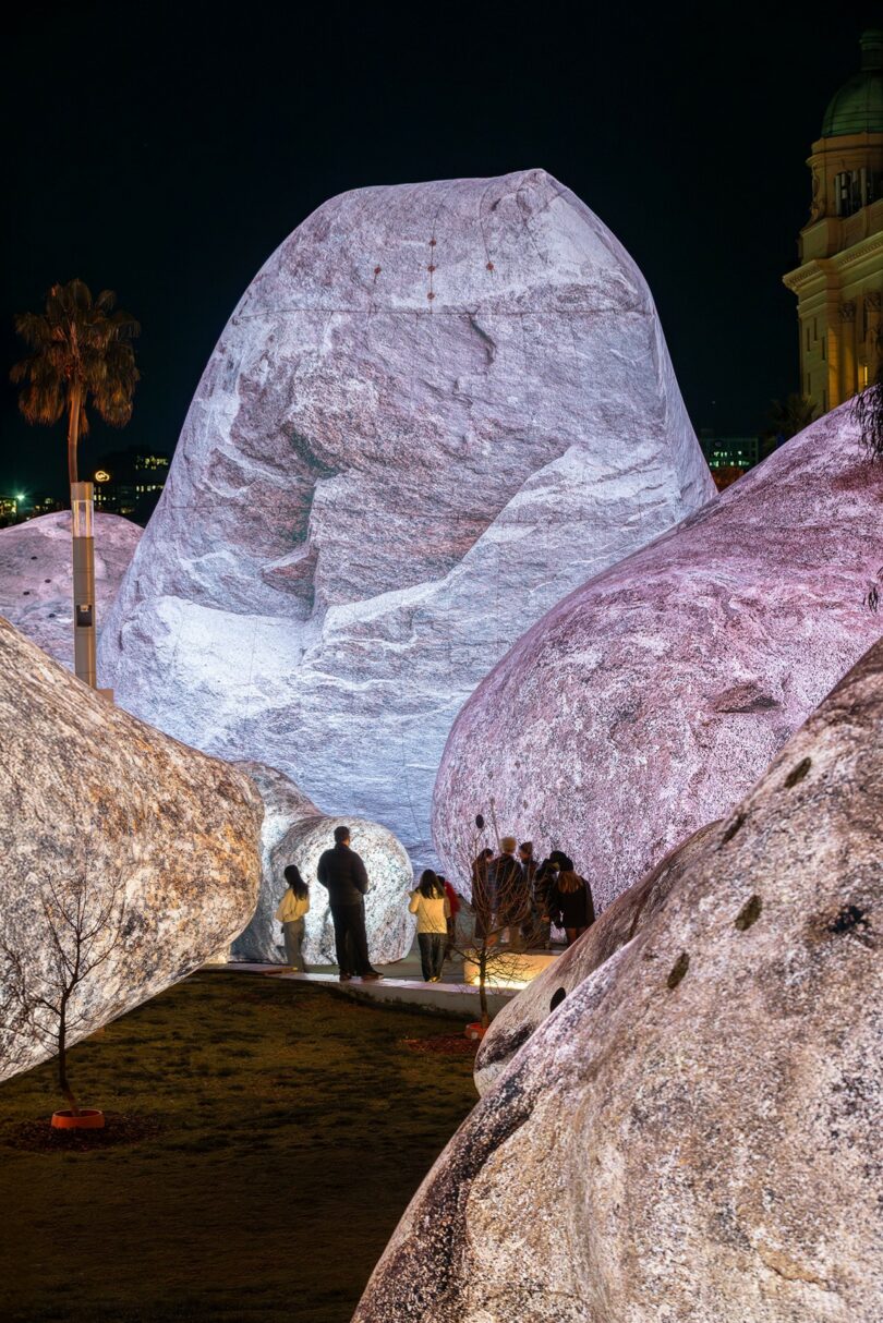 Large, illuminated rock-like structures outdoors at night with several people standing and walking among them; a palm tree and building are visible in the background