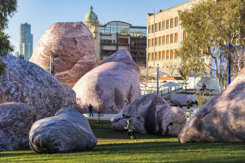 Large pink boulders scattered across a grassy urban park with people walking and running, modern and older buildings in the background