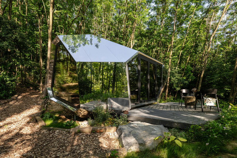 A modern, angular glass and mirrored cabin sits in a forest clearing, with a small wooden deck and two black chairs outside.