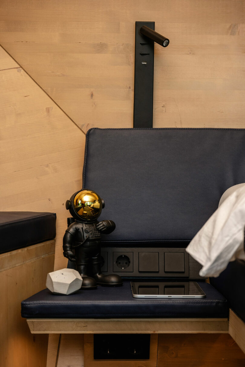 A small astronaut figurine with a gold helmet stands on a bench next to a white object, power outlets, and a bed with blue cushions in a wood-paneled room.
