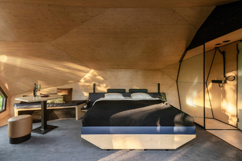 Modern bedroom with wood-paneled walls and ceiling, a large bed with dark bedding, a small dining table, and a minimalist open shower area. Natural light enters from a window.