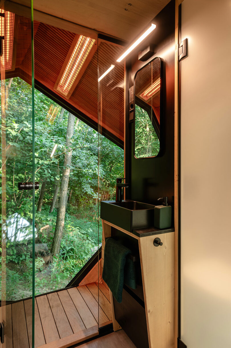 A modern bathroom with a black sink, wooden accents, glass walls, and a large window overlooking a lush forest.