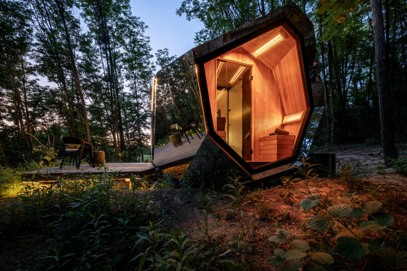 A small, modern cabin with angular design and illuminated wooden interior sits in a forested area at dusk, surrounded by trees and outdoor furniture.