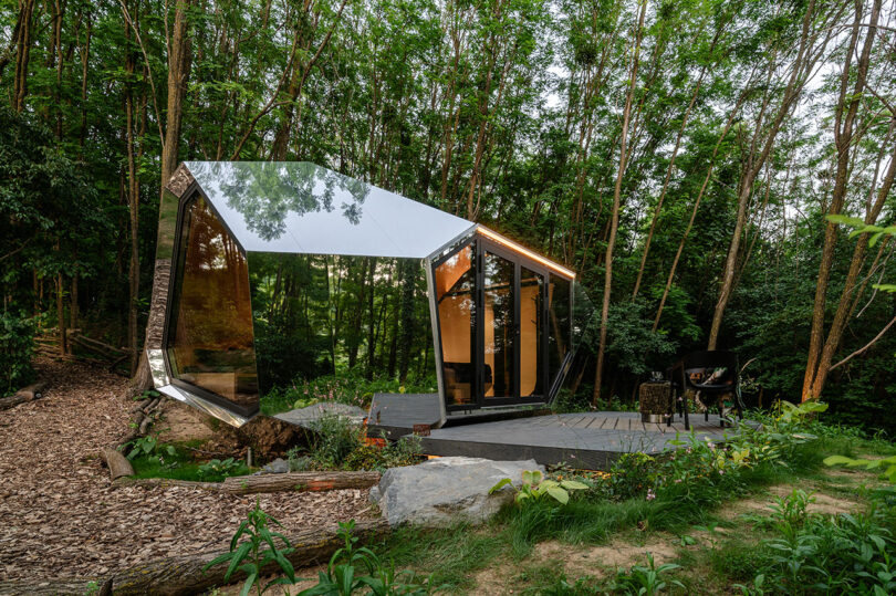 A modern, angular glass cabin with mirrored surfaces stands among trees in a forest, featuring a patio area with a grill on a wooden deck.