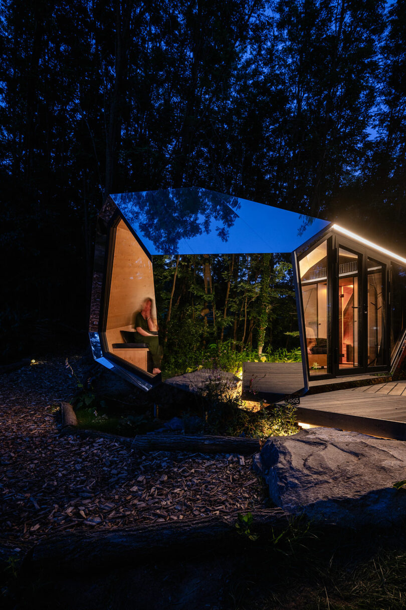 A modern, angular glass cabin with illuminated interior sits in a wooded area at dusk; a person is visible inside, seated near a window.