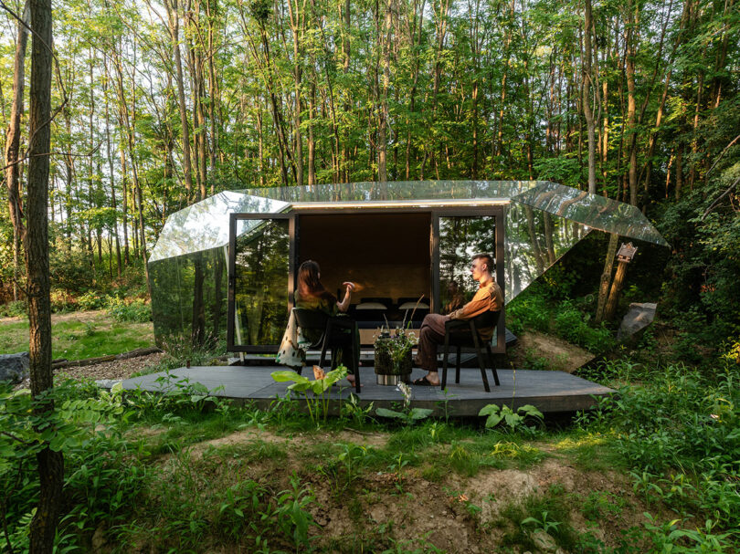 Two people sit and talk on a small wooden deck outside a modern, glass-paneled cabin surrounded by dense green forest.