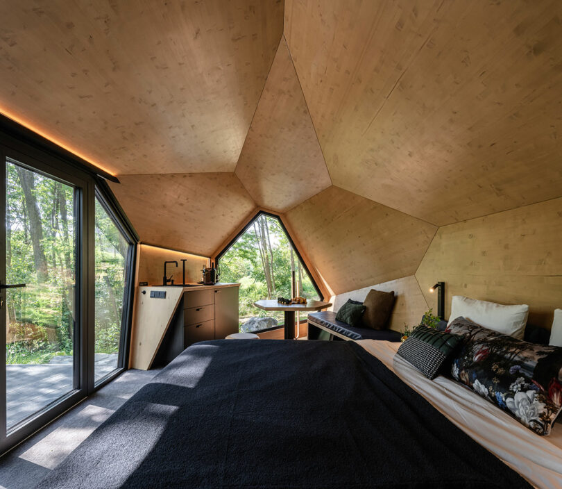 Modern bedroom with geometric wooden ceiling, large windows overlooking trees, a bed with dark bedding, a small kitchen area, and a table with two chairs. Natural light fills the space.