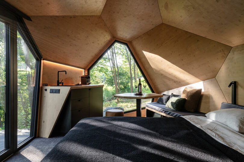 A modern cabin interior with geometric wood panel walls, large windows, a bed, a small kitchen area, and a table with seating, overlooking greenery outside.