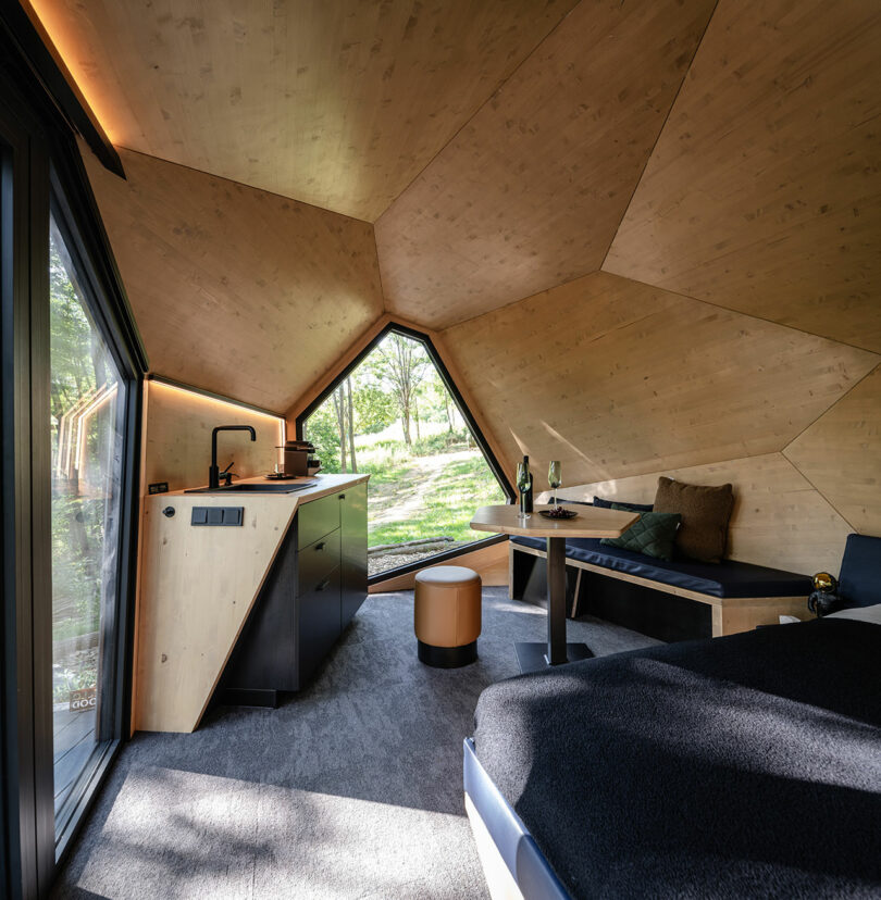 A modern, wood-paneled cabin interior with a geometric ceiling, kitchenette, bench seating, and large triangular window overlooking greenery.