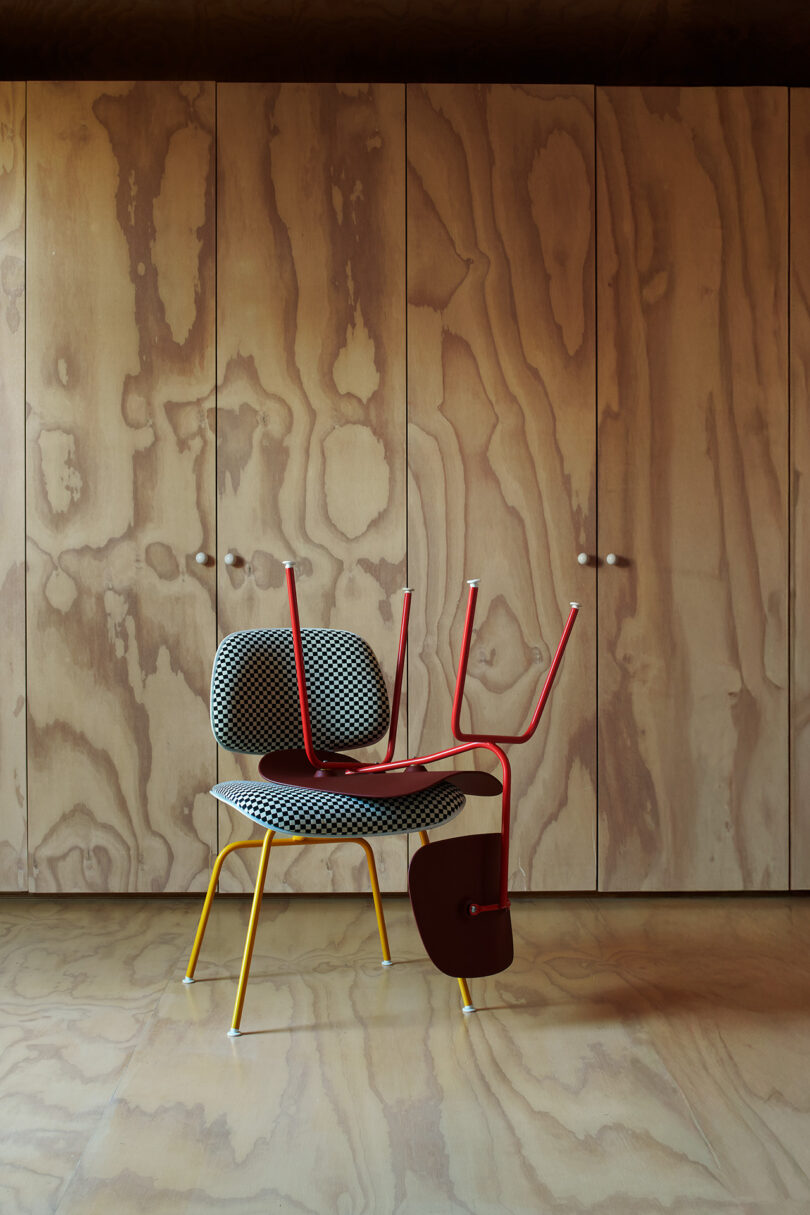 A patterned chair with yellow legs stands in front of wood-paneled doors, with a red chair placed upside down on top of it.