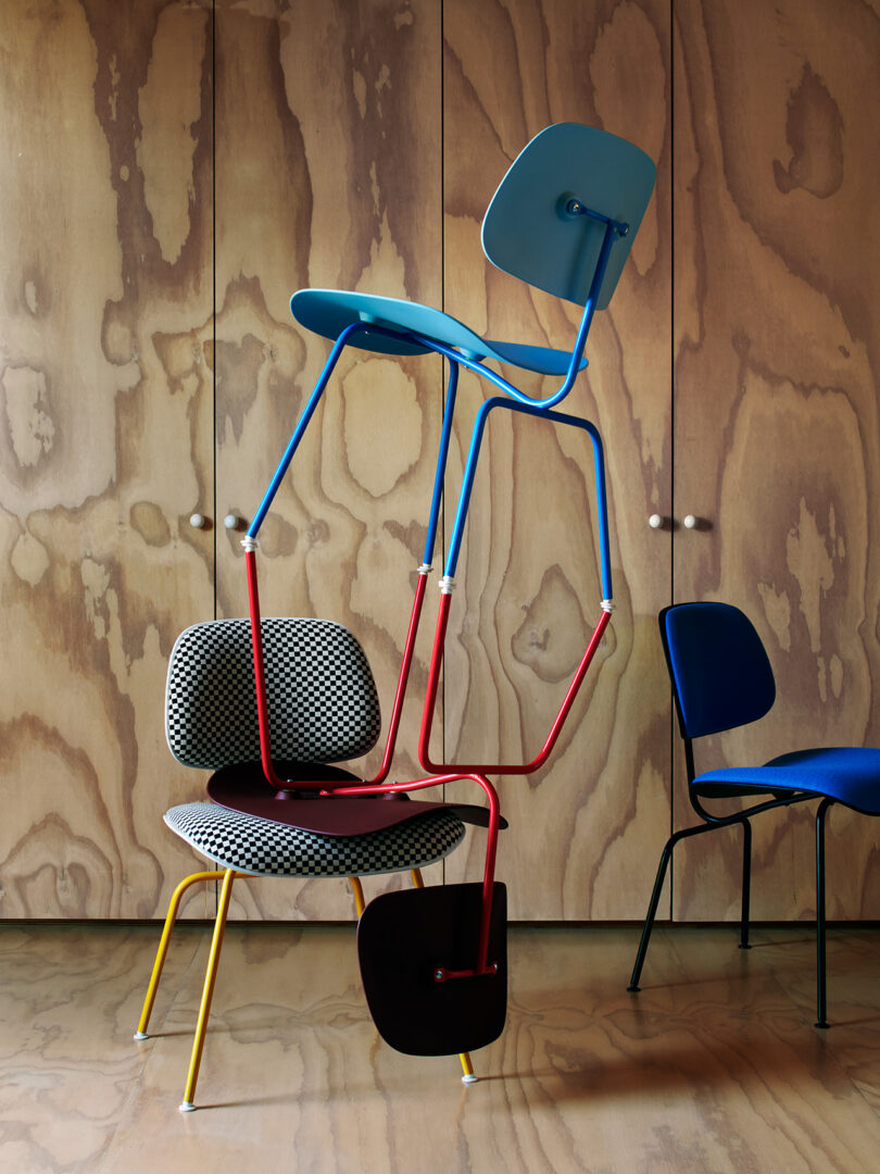 Three modern chairs with colorful metal legs are stacked in a precarious arrangement in front of a wooden paneled wall.