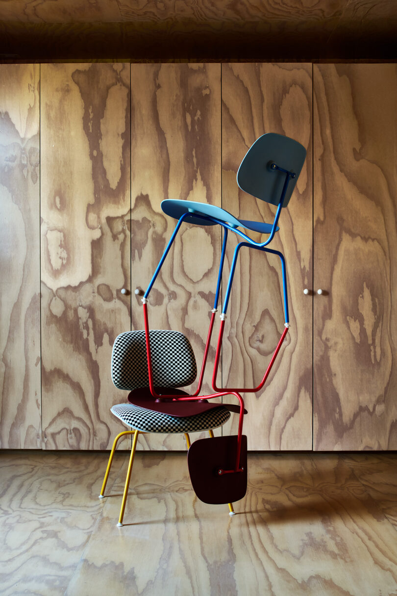A blue and red chair is balanced upside down on top of a patterned chair with yellow legs, set against a wooden wall with a distinctive grain pattern.