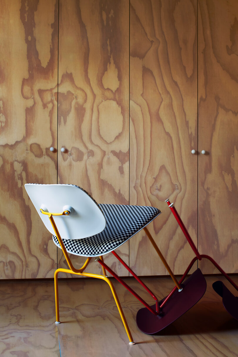 A modern chair with a yellow metal frame, white seat, and houndstooth-patterned cushion is positioned sideways on a wooden floor in front of plywood cabinets.