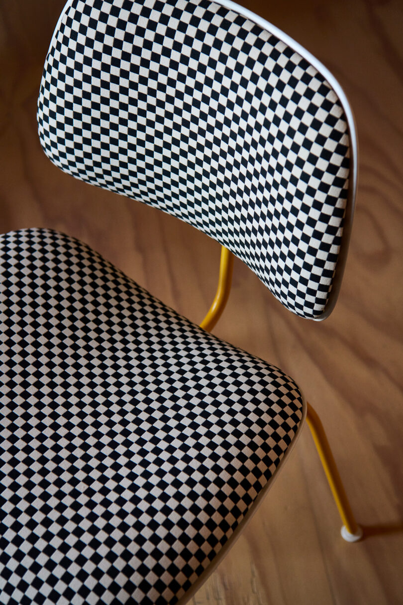 A close-up of a chair with a black-and-white checkerboard pattern on the seat and backrest, yellow legs, and a wooden floor background.